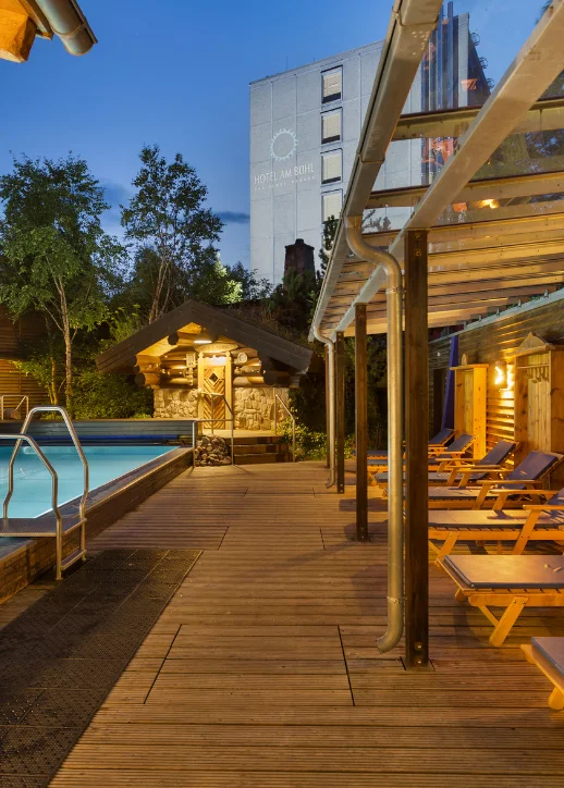 Holzdeck am Pool im Hotel am Bühl, Eibenstock, umgeben von Bäumen, bietet einen ruhigen Ort zum Entspannen und Genießen.
