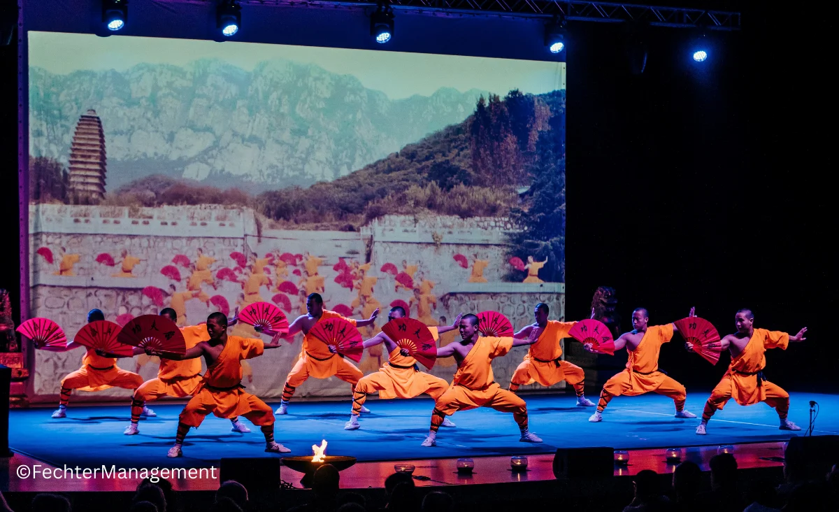 Mönche führen auf einer Bühne auf, während ein großer Bildschirm im Hintergrund sichtbar ist.