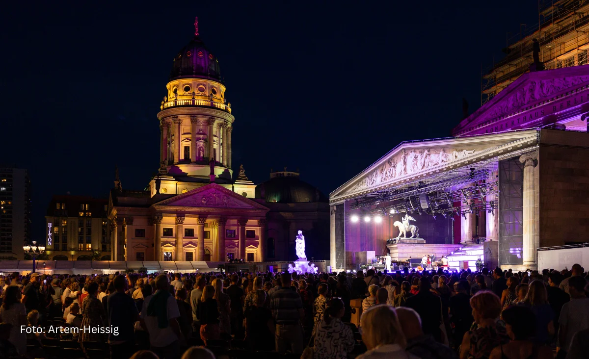 Bühnenaufnahme eines klassischen Open-Air-Konzerts mit einem Chor und einem Flügel im Hintergrund, davor ein Porträt eines Mannes mit offenem Hemdkragen.