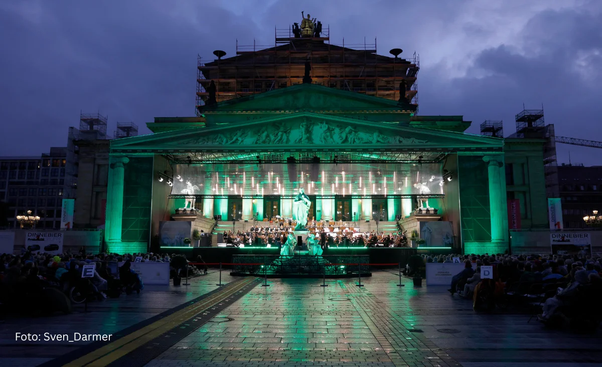 Bühnenaufnahme eines klassischen Open-Air-Konzerts mit einem Chor und einem Flügel im Hintergrund, davor ein Porträt eines Mannes mit offenem Hemdkragen.