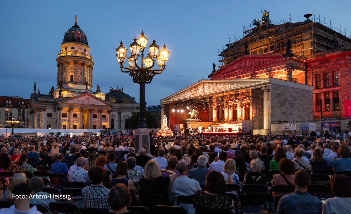 Bühnenaufnahme eines klassischen Open-Air-Konzerts mit einem Chor und einem Flügel im Hintergrund, davor ein Porträt eines Mannes mit offenem Hemdkragen.