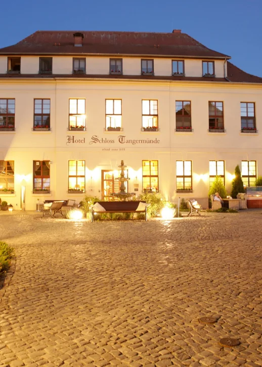 Beleuchtetes historisches Hotelgebäude mit gepflastertem Innenhof und Bäumen bei Abenddämmerung