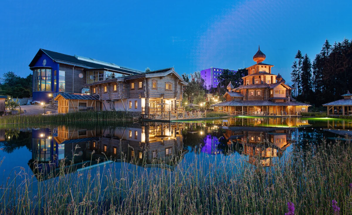 Holzgebäude mit traditioneller Architektur und beleuchteter Pagode spiegeln sich in einem ruhigen Teich im Badegärten Eibenstock bei Abenddämmerung