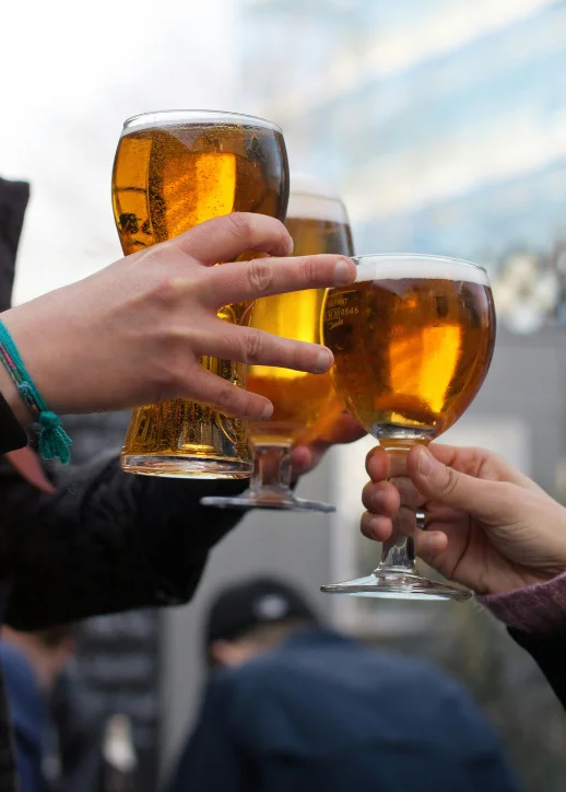 Drei Hände halten verschiedene Biergläser mit goldenem Bier und stoßen an. Im Hintergrund sind unscharfe Personen und moderne Gebäudefassaden zu erkennen. Die Szene spielt im Freien und vermittelt eine gesellige Atmosphäre, passend zum Bierfestival Berlin.