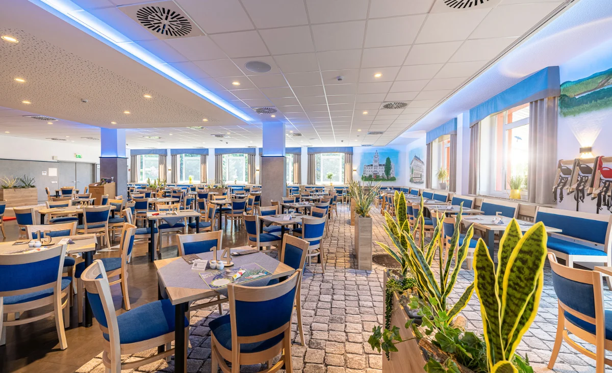 Großer, lichtdurchfluteter Speisesaal im Hotel Blaues Wunder Eibenstock mit zahlreichen gedeckten Tischen und blauen Stühlen. Die Einrichtung wirkt modern und freundlich, mit hellen Wänden, großen Fenstern und dekorativen Pflanzen. An den Wänden befinden sich Wandmalereien mit Landschaftsmotiven. Die Decke ist mit eingelassenen Leuchten und Lüftungselementen ausgestattet. Der Boden ist teilweise mit einem gemusterten Teppich ausgelegt, der an Kopfsteinpflaster erinnert.