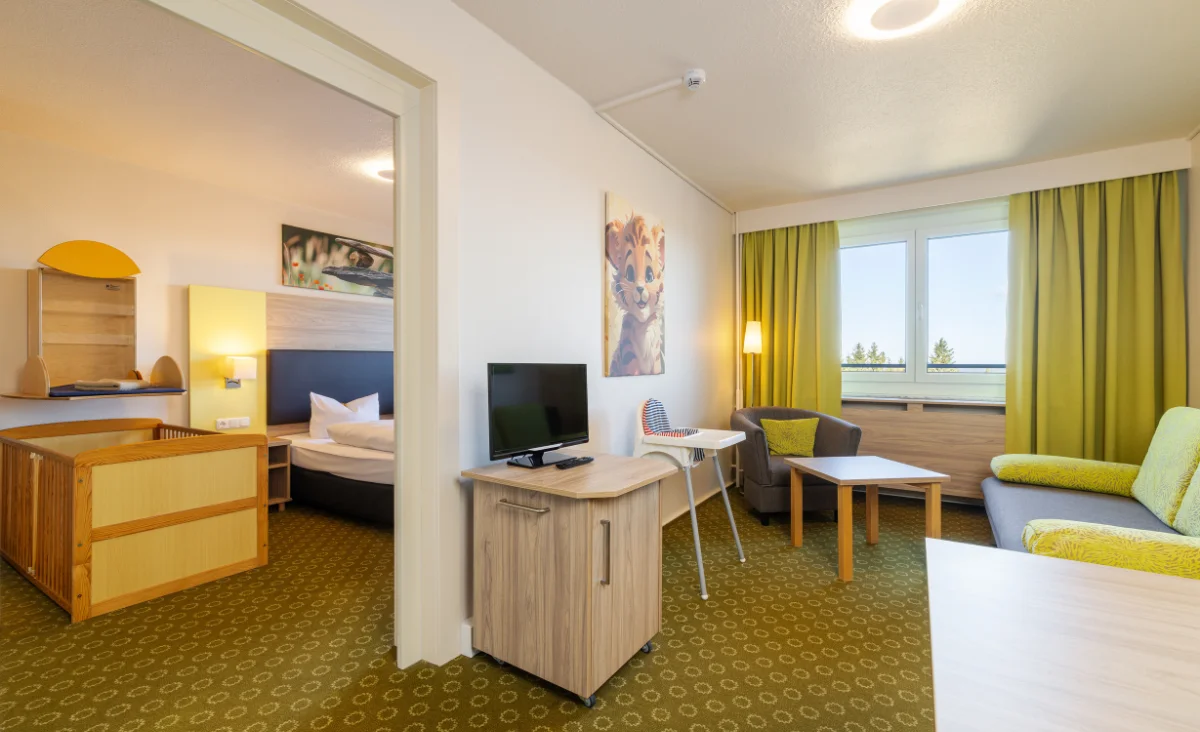 Im Vordergrund steht ein heller Holztisch mit einer kleinen Kommode, auf der ein Flachbildfernseher platziert ist. Rechts befindet sich ein grünes Sofa mit gelbem Polster, daneben ein grauer Sessel und ein weißer Couchtisch. Die Fenster sind mit gelb-grünen Vorhängen versehen und lassen Tageslicht herein. An der Wand hängt ein großformatiges Bild mit einem Tiermotiv. Im Hintergrund ist ein separates Schlafzimmer mit einem Doppelbett und einem Kinderbett zu sehen. Die Einrichtung wirkt modern und freundlich, die Teppichböden sind mit einem kreisförmigen Muster gestaltet.
