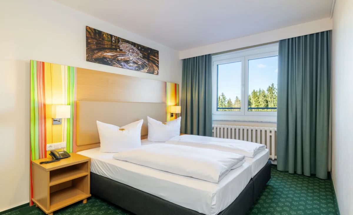 Doppelzimmer im Hotel Blaues Wunder Eibenstock mit zwei nebeneinanderstehenden Betten, weißer Bettwäsche und zwei großen Kissen. Die Wand hinter dem Bett ist mit einem farbigen Streifenmuster in Gelb-, Grün- und Orangetönen gestaltet. Über dem Betthaupt hängt ein rechteckiges Bild mit einem abstrakten Motiv. Links neben dem Bett steht ein Nachttisch mit Telefon und Notizblock, an der Wand sind zwei Wandleuchten angebracht. Vor dem Fenster mit hellblauen Vorhängen ist ein Heizkörper zu sehen. Der Teppichboden ist grün mit dezentem Muster. Durch das Fenster fällt Tageslicht und es ist ein Ausblick auf Bäume erkennbar.