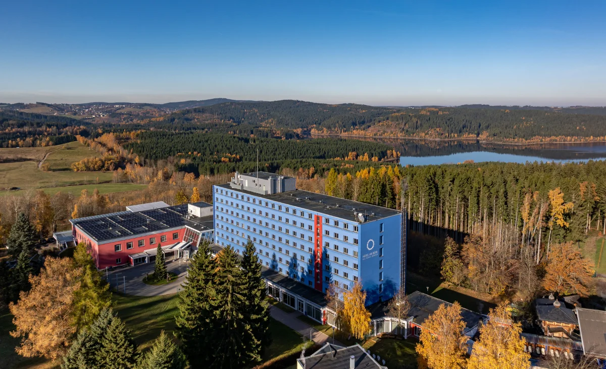 Das Hotel Blaues Wunder Eibenstock steht im Vordergrund einer herbstlichen Landschaft. Das Hauptgebäude ist mehrstöckig, in einem hellen Blauton gestrichen und besitzt eine markante rote vertikale Linie an der Fassade. Rechts davon erstreckt sich ein großer Nadelwald, dahinter liegt ein See. Im Hintergrund sind sanfte Hügel und weitere Waldflächen zu sehen. Die Bäume im Umfeld des Hotels zeigen gelbe und orangefarbene Herbstfärbung. Der Himmel ist wolkenlos und blau.