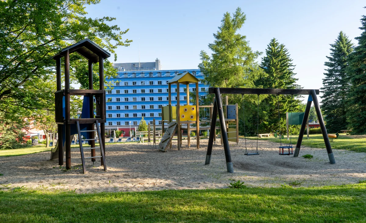 Im Vordergrund befindet sich ein Spielplatz mit verschiedenen Holzkonstruktionen, darunter ein Kletterturm, eine Rutsche, Schaukeln und Kletterelemente. Der Boden ist mit hellem Sand bedeckt. Im Hintergrund steht das Hotel Blaues Wunder Eibenstock, ein mehrstöckiges Gebäude mit blauer Fassade und zahlreichen Fenstern. Der Bereich ist von Rasenflächen und hohen Nadelbäumen umgeben, die für eine grüne, naturnahe Umgebung sorgen. Die Szene ist von Tageslicht erhellt.