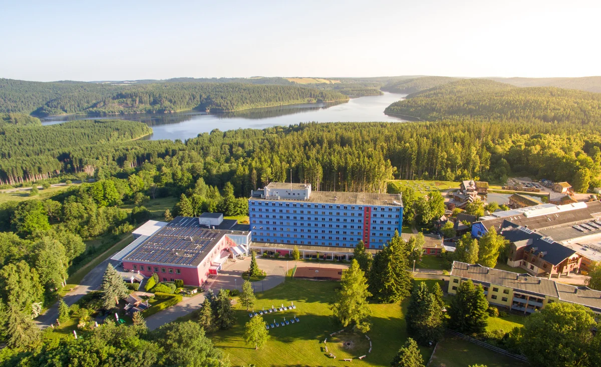 Das Hotel Blaues Wunder Eibenstock steht im Vordergrund einer Luftaufnahme. Das mehrstöckige Gebäude ist blau und hebt sich deutlich von den umliegenden, niedrigeren Gebäuden ab. Im Hintergrund erstreckt sich eine waldreiche Hügellandschaft mit einem großen, geschwungenen See. Die Umgebung ist von dichtem Wald und weitläufigen Grünflächen geprägt. Auf der Wiese vor dem Hotel sind mehrere Sitzgelegenheiten angeordnet. Die Aufnahme vermittelt einen Eindruck von ruhiger, naturnaher Lage.