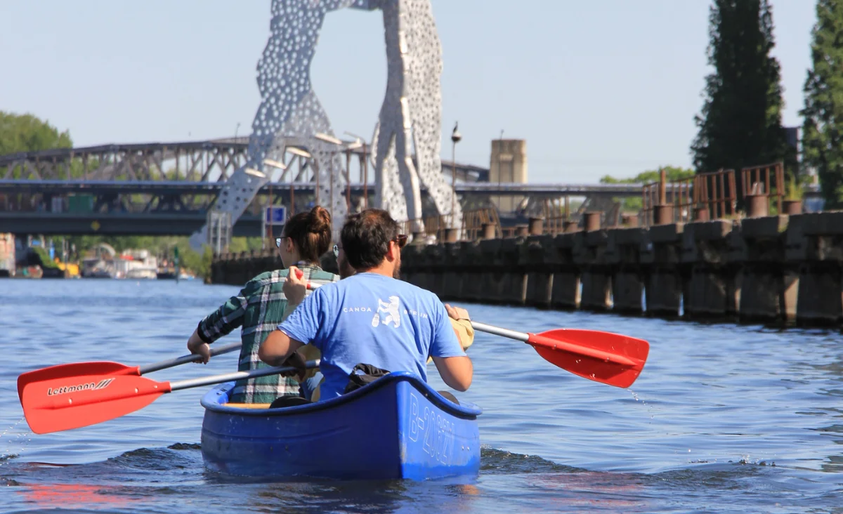 Zwei Personen paddeln in einem blauen Kanu auf einem Fluss mit der Skulptur "Molecule Man" und einer Brücke im Hintergrund