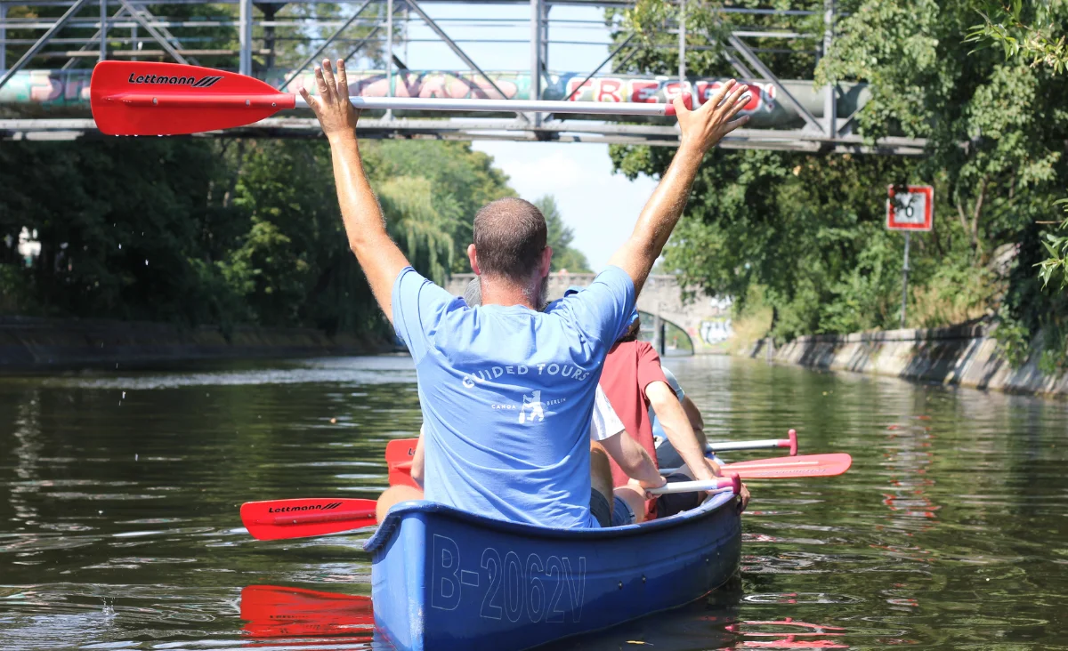 Person in blauem T-Shirt mit Aufschrift 'GUIDED TOUR' sitzt in blauem Kanu und hält rotes Paddel über den Kopf auf einem Fluss unter einer Brücke. Die Szene vermittelt den Eindruck einer geführten Sightseeing-Gruppen-Tour auf dem Wasser.