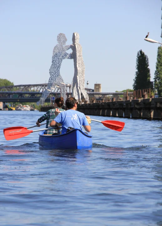 Zwei Personen paddeln in einem blauen Kanu auf einem Fluss mit der Skulptur "Molecule Man" und einer Brücke im Hintergrund