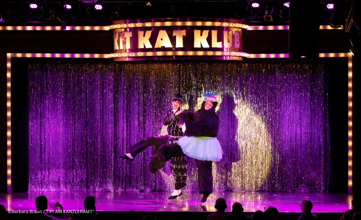 Zwei Personen stehen auf einer Bühne des Musicals »Cabaret Berlin! – das Berlin Musical« vor einem Vorhang aus silbernen Lamellen. Über ihnen leuchtet ein Schild mit der Aufschrift »KIT KAT KLUB« in großen, beleuchteten Buchstaben.
