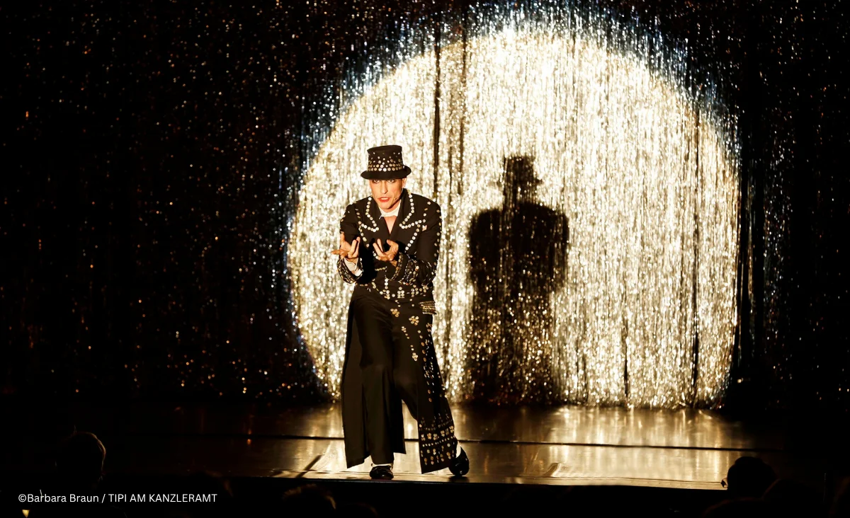 Ein Künstler auf der Bühne in einem glitzernden Outfit, umgeben von funkelndem Hintergrund und dramatischer Beleuchtung.