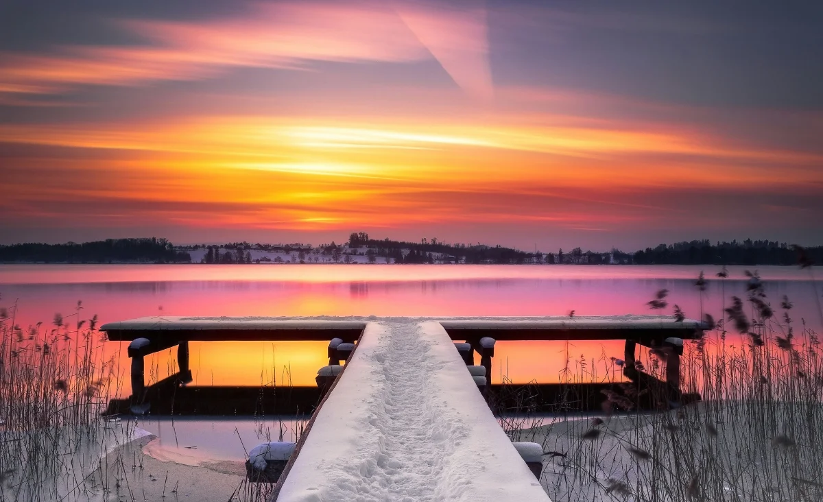 Steg mit Schnee über einem ruhigen See bei Sonnenuntergang mit orange-rotem Himmel und Schilf am Ufer.