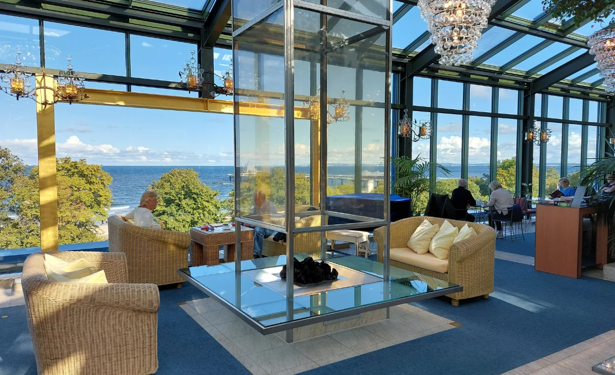 Lounge mit Korbsesseln und Sofa in einem verglasten Raum mit Blick auf die Ostsee und Bäume aus dem Hotel Kaiserhof Heringsdorf