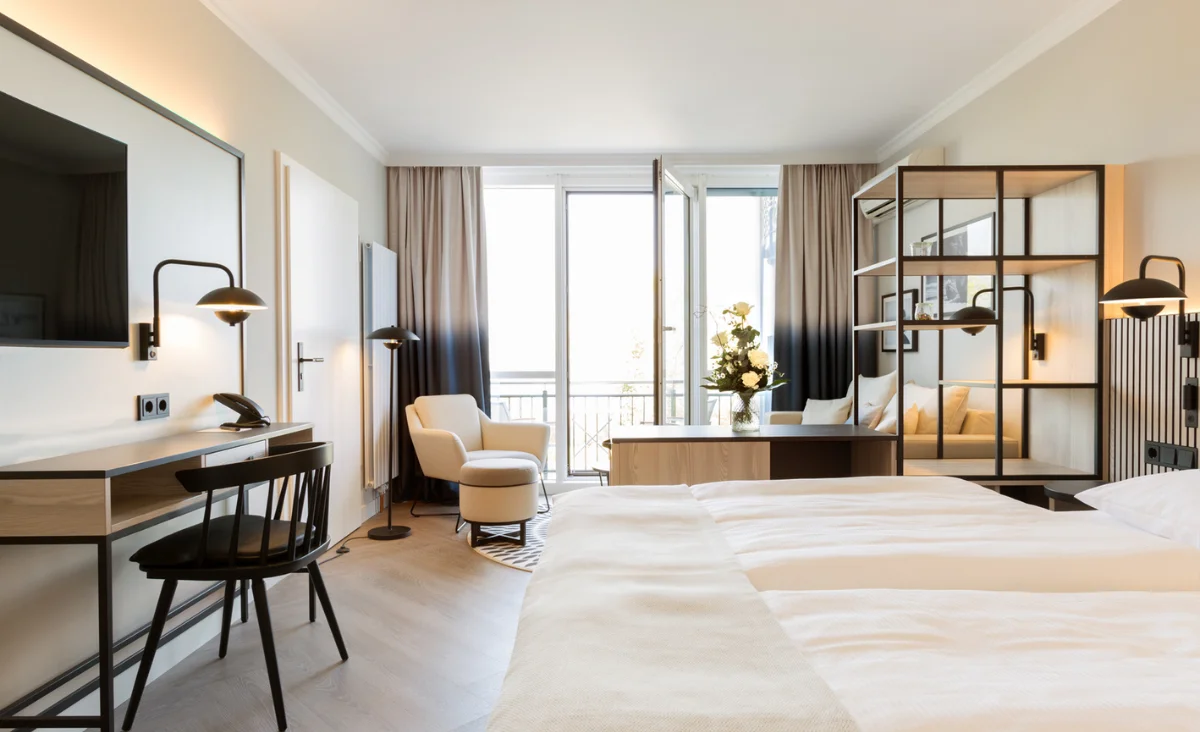 Helles Hotelzimmer mit Sofa, Sessel, rundem Tisch, offenem Balkonfenster und Blick auf Bäume aus dem Hotel Kaiserhof Heringsdorf