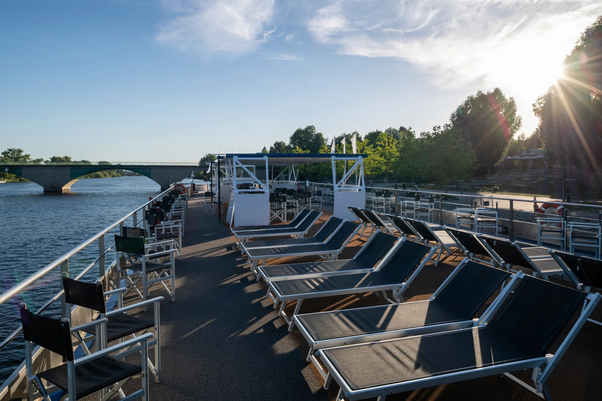 Sonnendurchflutetes Sonnendeck eines Flusskreuzfahrtschiffs mit Reihen von Liegestühlen und Stühlen neben einem Fluss und einer Brücke im Hintergrund