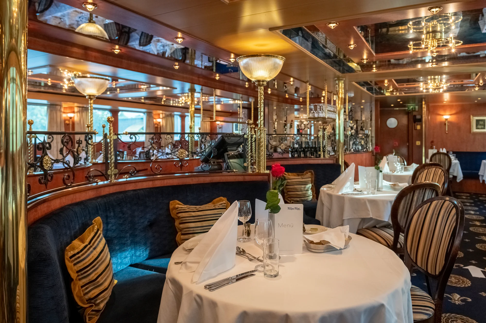 Elegantes Restaurant an Bord eines Flusskreuzfahrtschiffs mit runden, weiß eingedeckten Tischen, auf denen Menükarten, Weingläser, Besteck und eine einzelne rote Rose in einer Vase stehen. Gepolsterte Sitzbänke mit gestreiften Kissen und klassische Stühle mit gestreiften Bezügen umgeben die Tische. Die Decke ist verspiegelt und mit zahlreichen kleinen Leuchten sowie goldfarbenen Elementen verziert. Im Hintergrund sind dekorative Geländer und Spiegelwände zu sehen. Die Atmosphäre wirkt festlich und einladend, passend zur Adventsreise auf der Havel und Oder mit Thurgau Travel.