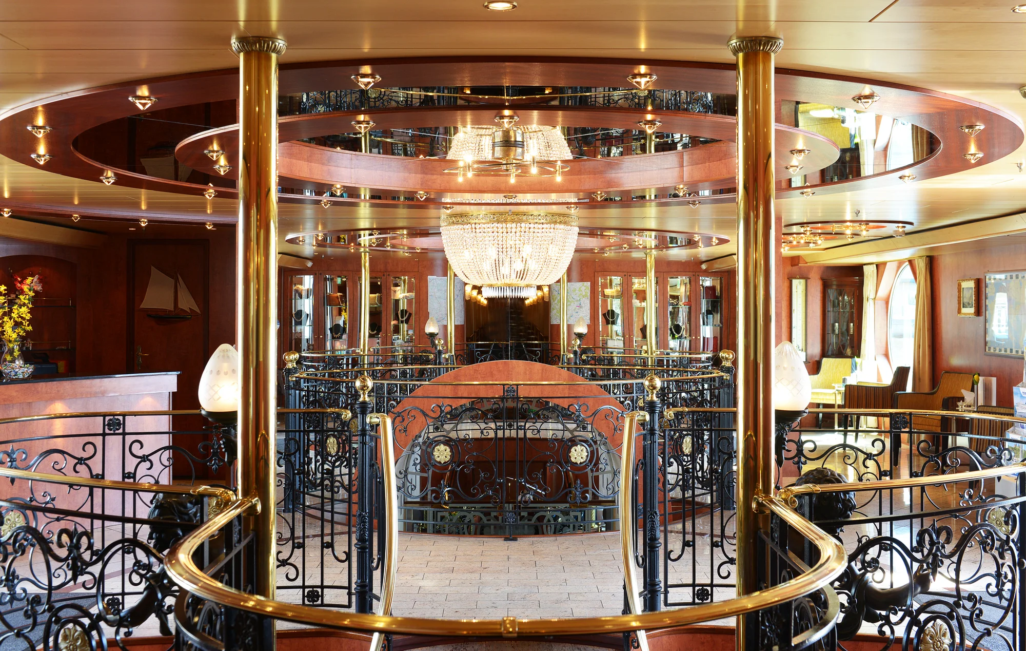 Zentraler Blick in den großzügigen, elegant gestalteten Salon eines Flusskreuzfahrtschiffs der Marke Thurgau Travel. Im Mittelpunkt hängt ein großer, funkelnder Kronleuchter unter einer abgehängten Decke mit Spiegel- und Holzelementen. Mehrere goldfarbene Säulen und kunstvoll verzierte, schwarze Geländer mit floralen Mustern umrahmen den offenen Bereich. Im Hintergrund sind eine Rezeption mit Blumendekoration und große Fenster mit hellen Vorhängen zu sehen. Die Einrichtung wirkt hochwertig und auf Komfort ausgelegt.