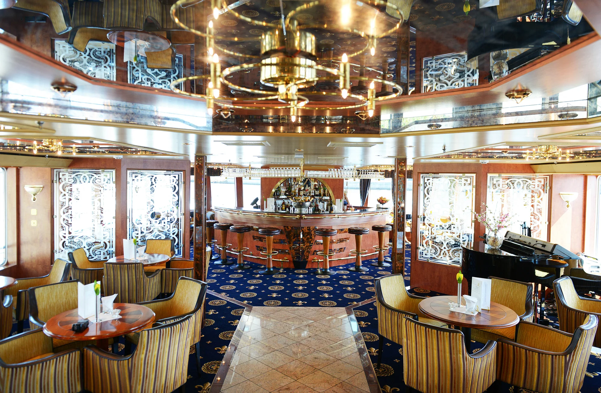Eleganter Innenraum eines Flusskreuzfahrtschiffs der Marke Thurgau Travel mit zentraler Bar, runder Deckenbeleuchtung und spiegelnder Decke. Die Einrichtung umfasst runde Holztische mit gestreiften Polsterstühlen, auf denen weiße Servietten und einzelne Blumen in Vasen stehen. Der Boden ist mit einem blauen Teppich mit goldfarbenem Muster und einem hellen Marmormittelgang ausgelegt. Seitlich sind dekorative Glaselemente mit floralen Mustern zu sehen. Rechts steht ein schwarzer Flügel.