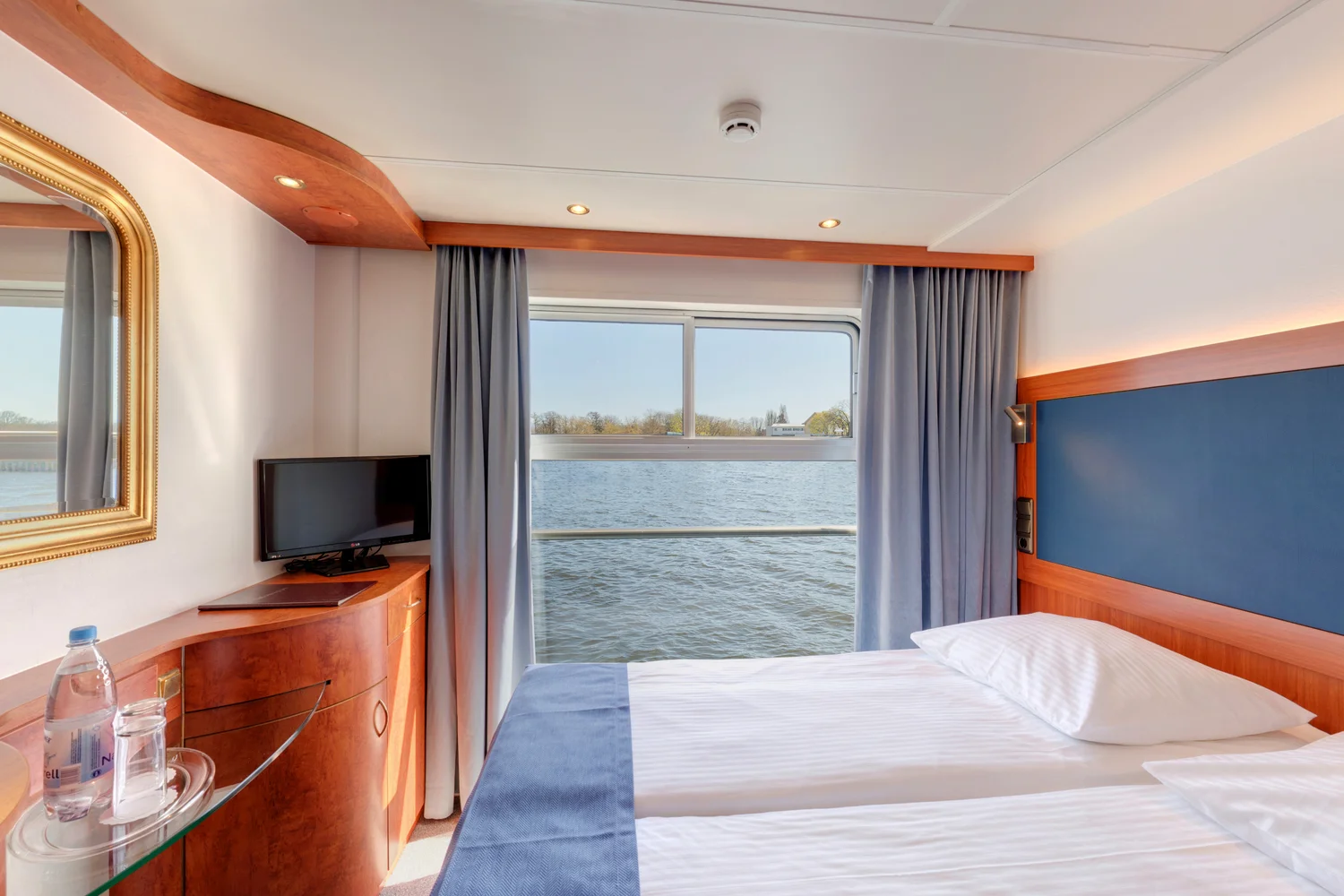 Helles Kabinenzimmer mit zwei nebeneinanderstehenden Einzelbetten, weißer Bettwäsche und blauer Tagesdecke. An der rechten Wand befindet sich ein großes Fenster mit Blick auf einen Fluss und das gegenüberliegende Ufer mit Bäumen. Links steht ein kleiner Flachbildfernseher auf einem Holzschrank, daneben eine Glasablage mit einer Wasserflasche und einem Glas. Die Einrichtung ist in warmen Holztönen gehalten, die Vorhänge sind hellblau und grau. Die Szene vermittelt den Eindruck einer komfortablen Unterkunft auf einem Flusskreuzfahrtschiff, passend zur Flusskreuzfahrt „Advent auf Havel & Oder“ von Thurgau Travel.
