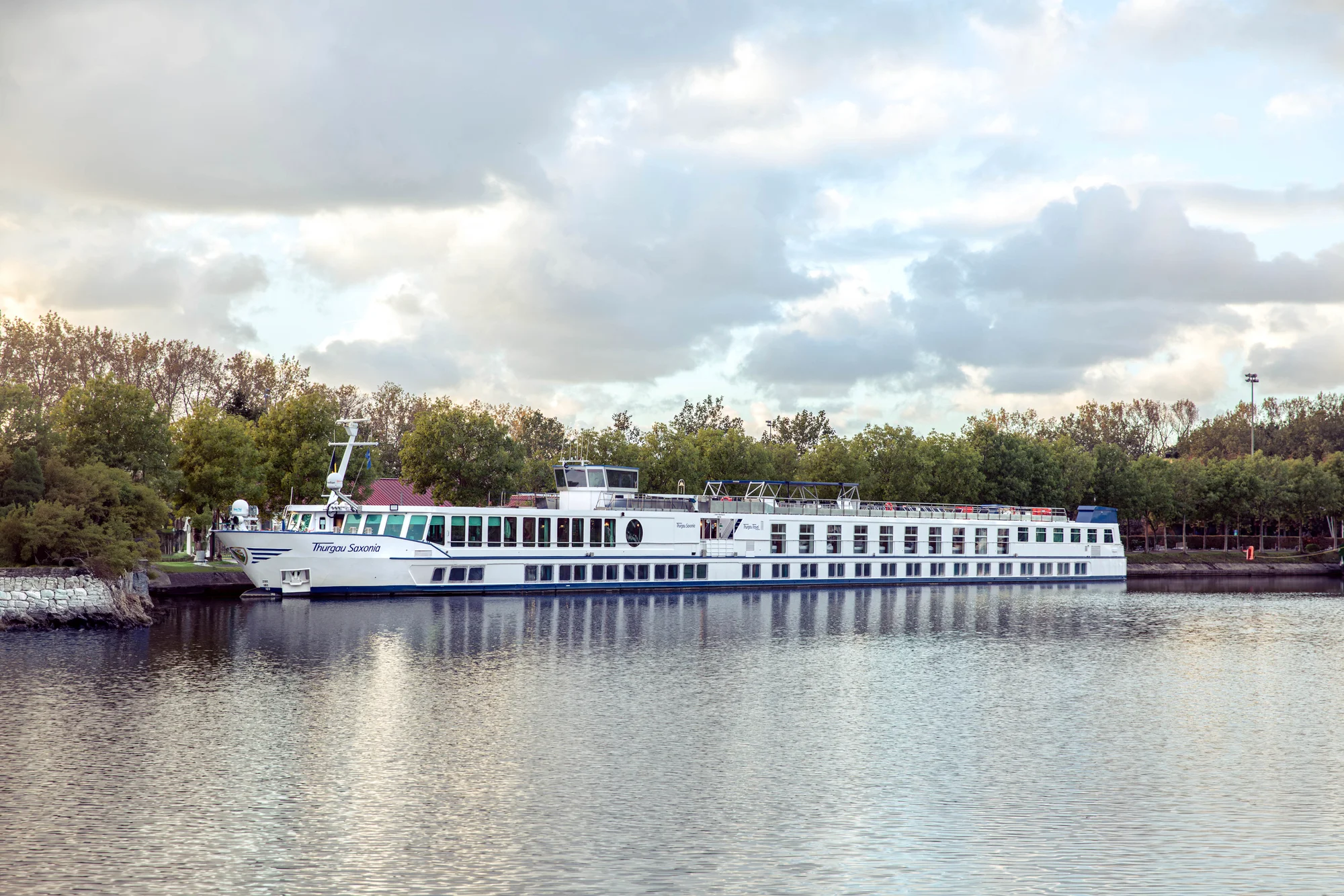 Das Flusskreuzfahrtschiff »Thurgau Saxonia« liegt am Ufer eines ruhigen Flusses vor einer Baumreihe. Das Schiff ist langgestreckt, weiß lackiert und trägt einen blauen Streifen entlang des Rumpfes. Auf dem Oberdeck befinden sich Sitzgelegenheiten und Sonnenschutz. Die Szenerie ist von bewölktem Himmel und sanftem Licht geprägt. Die Aufnahme vermittelt eine ruhige, entspannte Atmosphäre, passend zur Flusskreuzfahrt »Advent auf Havel & Oder« von Thurgau Travel.