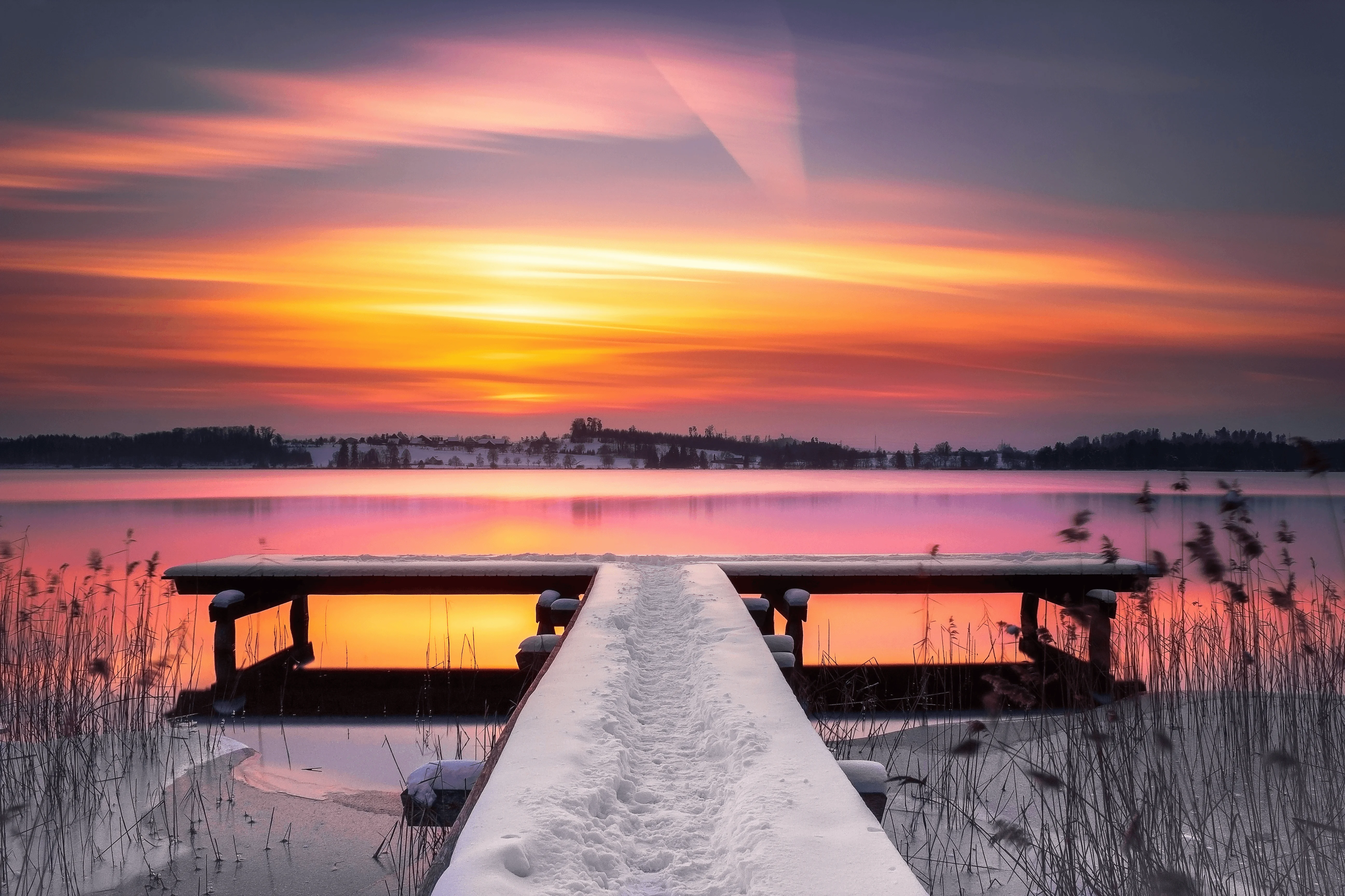 Steg mit Schnee über einem ruhigen See bei Sonnenuntergang mit orange-rotem Himmel und Schilf am Ufer