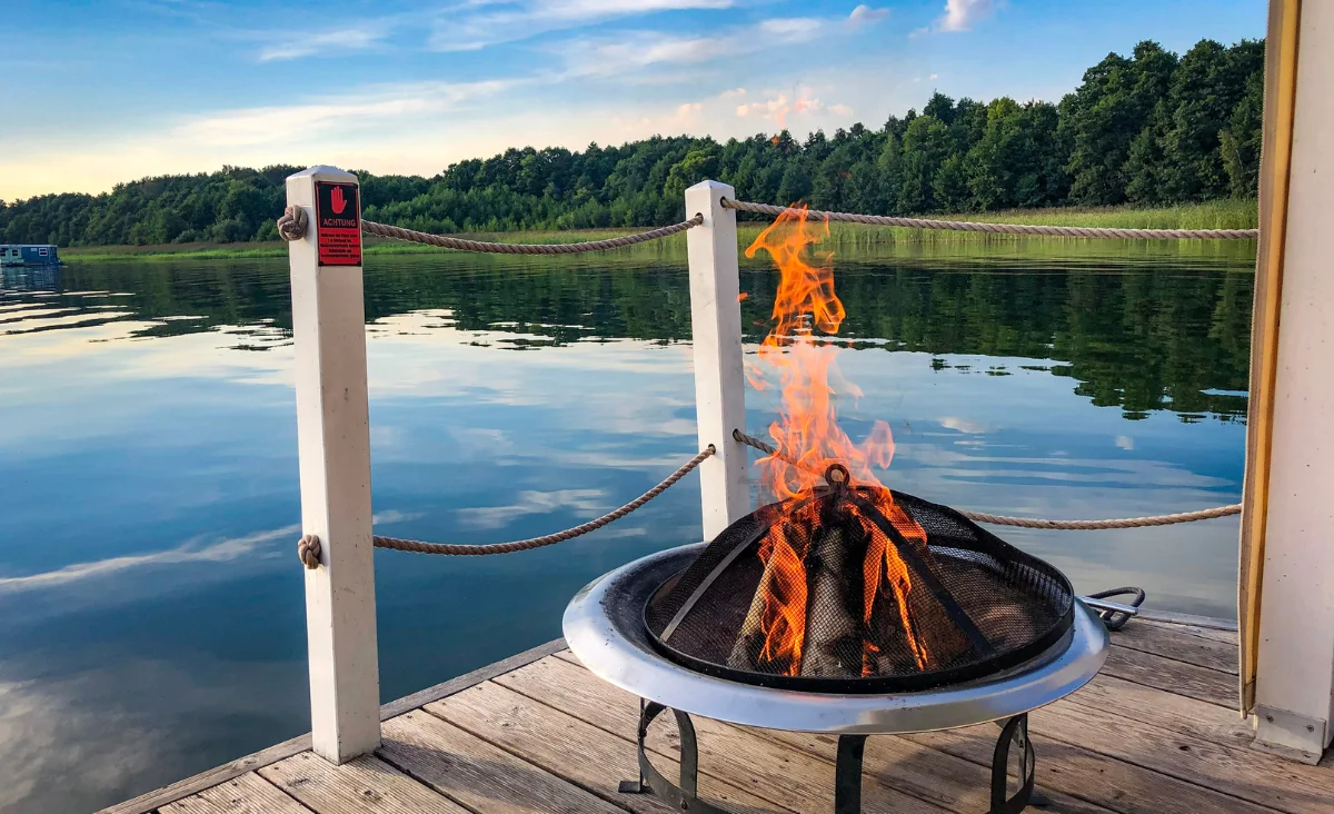 Brennendes Feuer in einer Feuerschale auf einem Holzsteg mit Blick auf einen ruhigen See und bewaldetes Ufer im Hintergrund