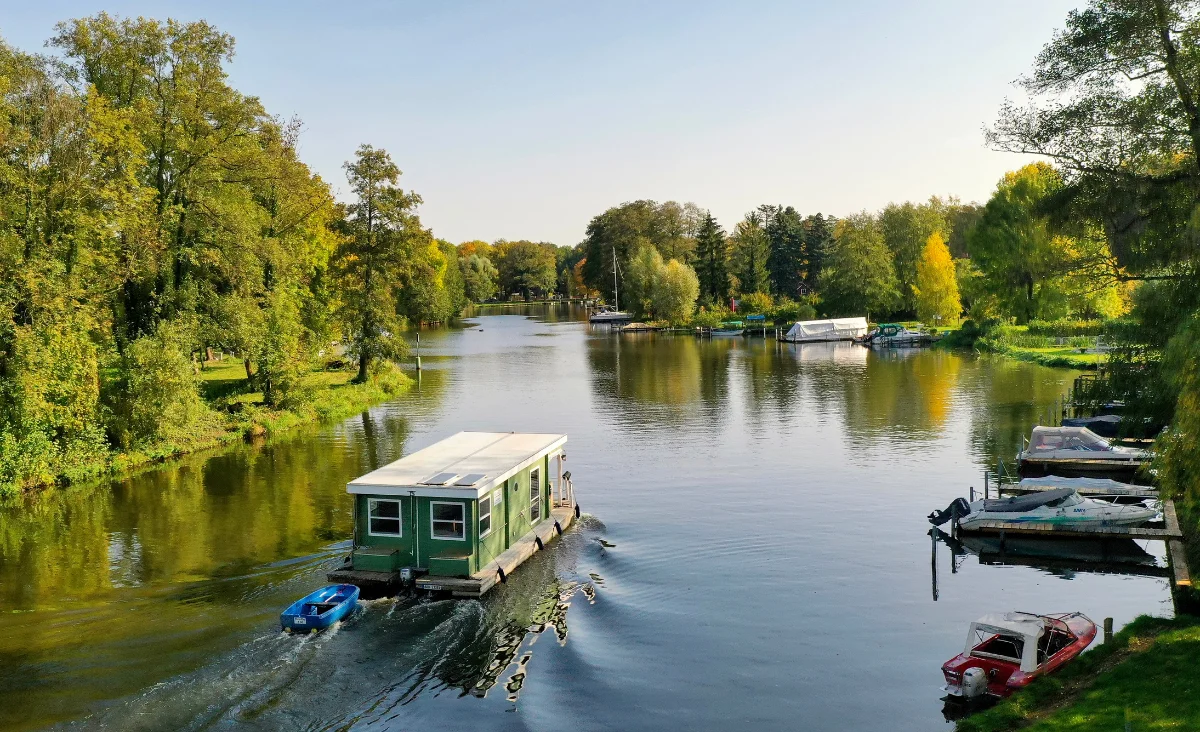 Hausboot fährt auf einem ruhigen Fluss umgeben von grünen Bäumen und mehreren angelegten Booten