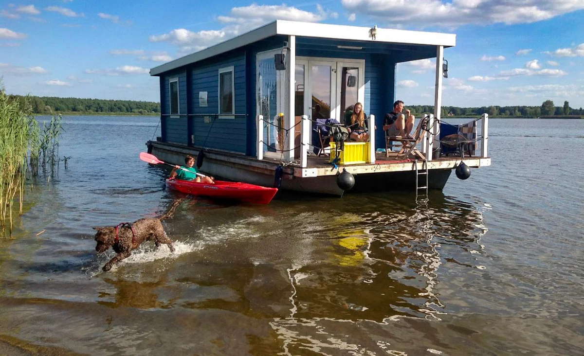 Hausboot auf ruhigem See mit zwei Personen auf der Terrasse, einem Hund im Wasser und einer Person im roten Kajak
