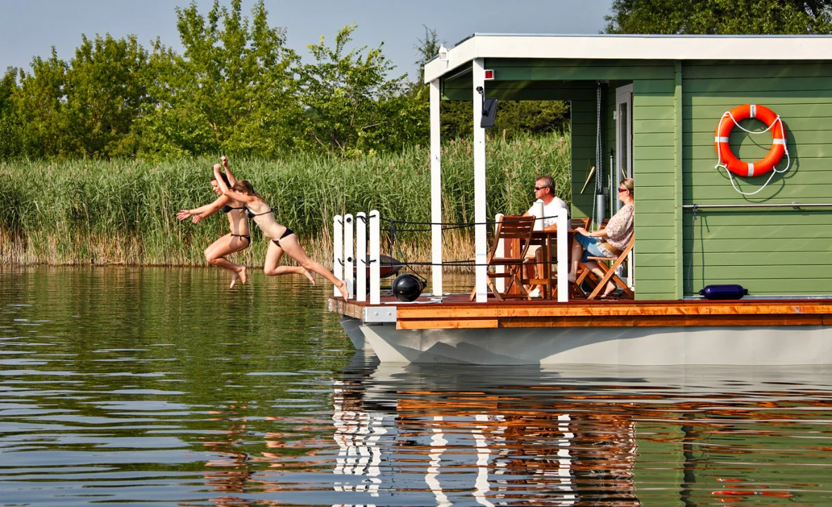 Zwei Frauen springen vom Hausboot ins Wasser während zwei Männer auf dem Hausboot auf Stühlen sitzen und zuschauen