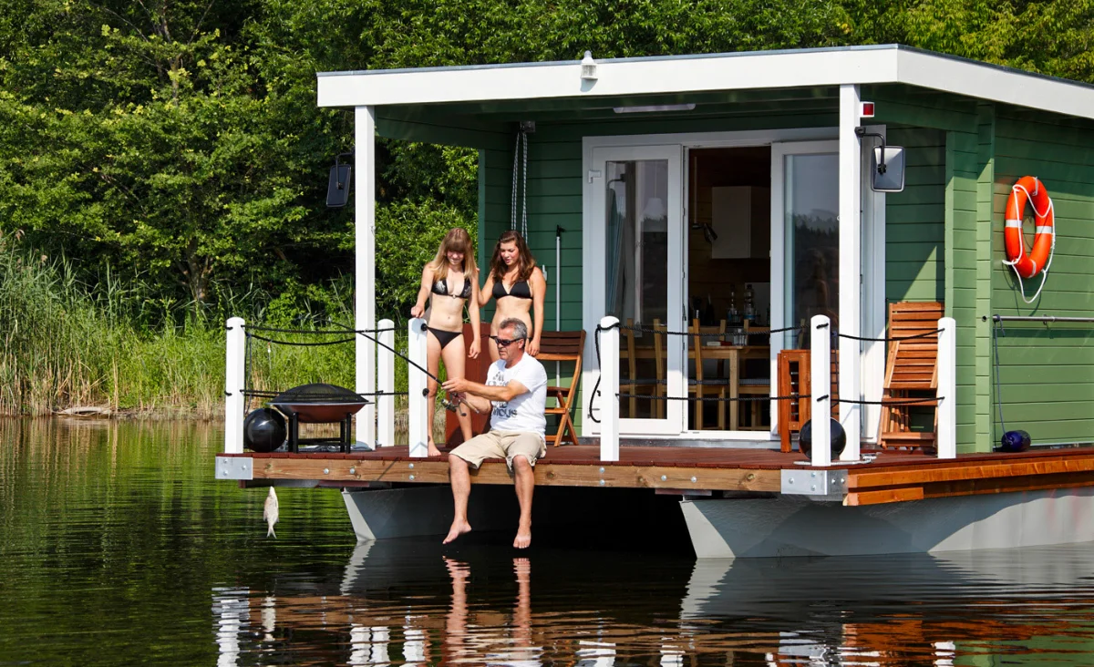 Hausboot auf ruhigem Wasser mit zwei Frauen in Bikinis und einem Mann, der angelt
