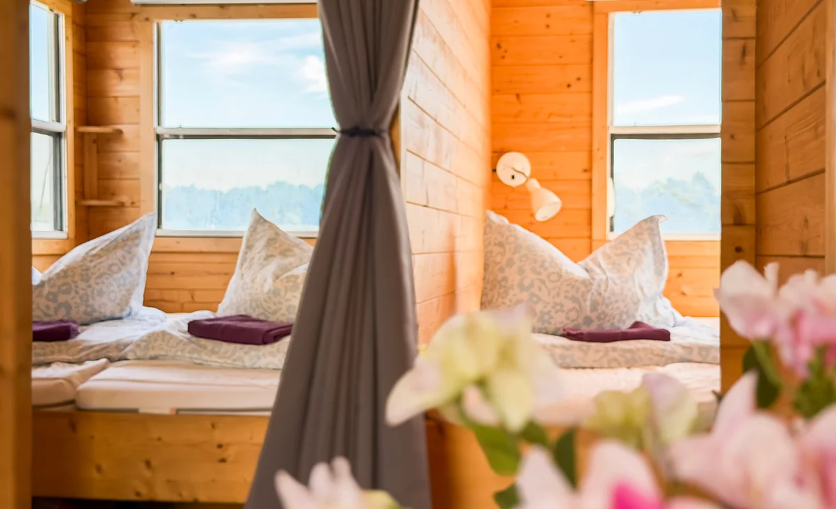Zwei Betten mit gemusterten Kissen und dunkelroten Handtüchern in einem hellen Holzraum, getrennt durch einen grauen Vorhang, im Vordergrund unscharfe Blumen