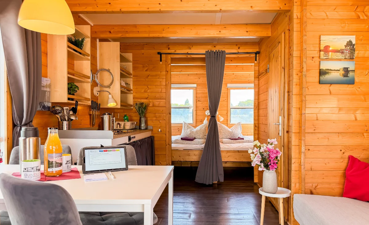 Innenraum eines Holzhausboots mit Esstisch, Laptop, Küchenzeile, Vorhang und Bett im Hintergrund