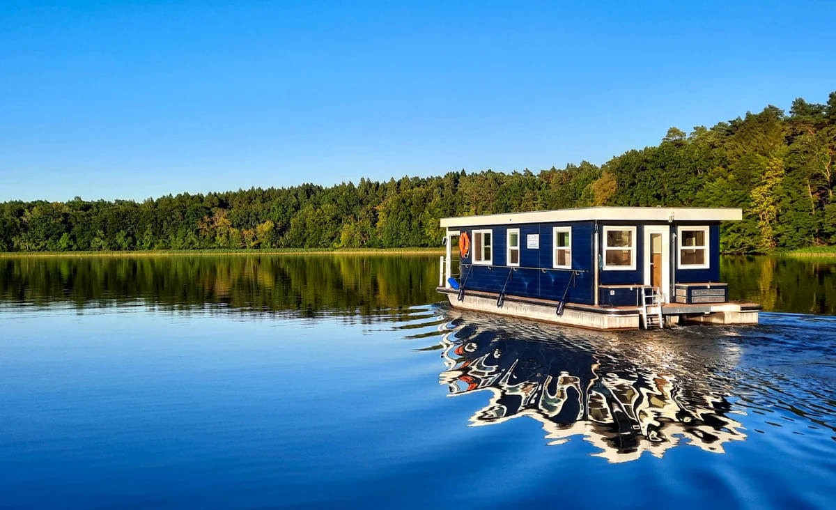 Hausboot mit rechteckigem Aufbau und mehreren Fenstern fährt auf ruhigem See vor bewaldetem Ufer unter klarem Himmel