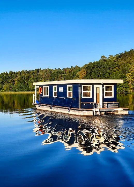 Hausboot mit rechteckigem Aufbau und mehreren Fenstern fährt auf ruhigem See vor bewaldetem Ufer unter klarem Himmel