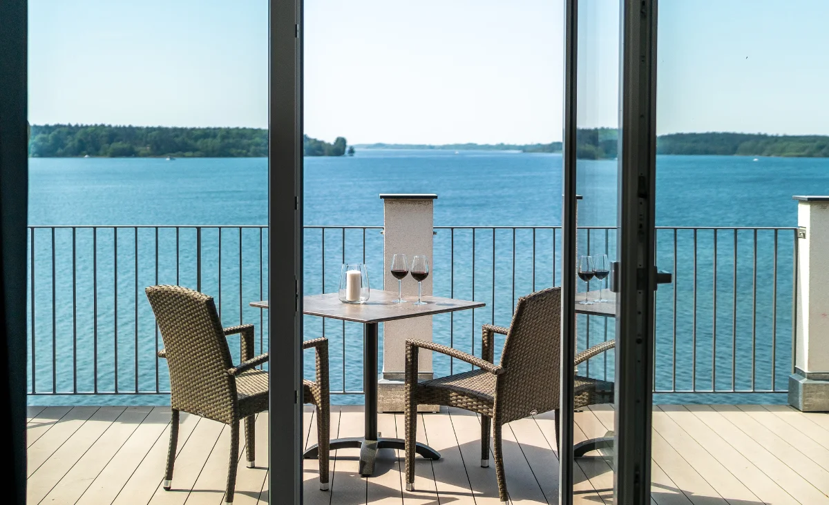 Balkon mit zwei geflochtenen Stühlen und einem kleinen Tisch mit zwei Weingläsern und einer Kerze mit Blick auf einen großen See und bewaldete Ufer im Hintergrund