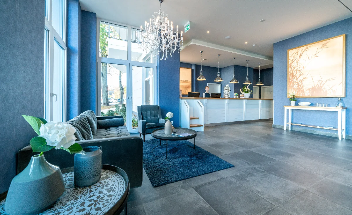 Heller Empfangsbereich im Aparthotel Müritzpalais mit blauem Sofa, Sesseln, Teppich, runder Tisch mit Vasen und Kronleuchter über der Rezeption.