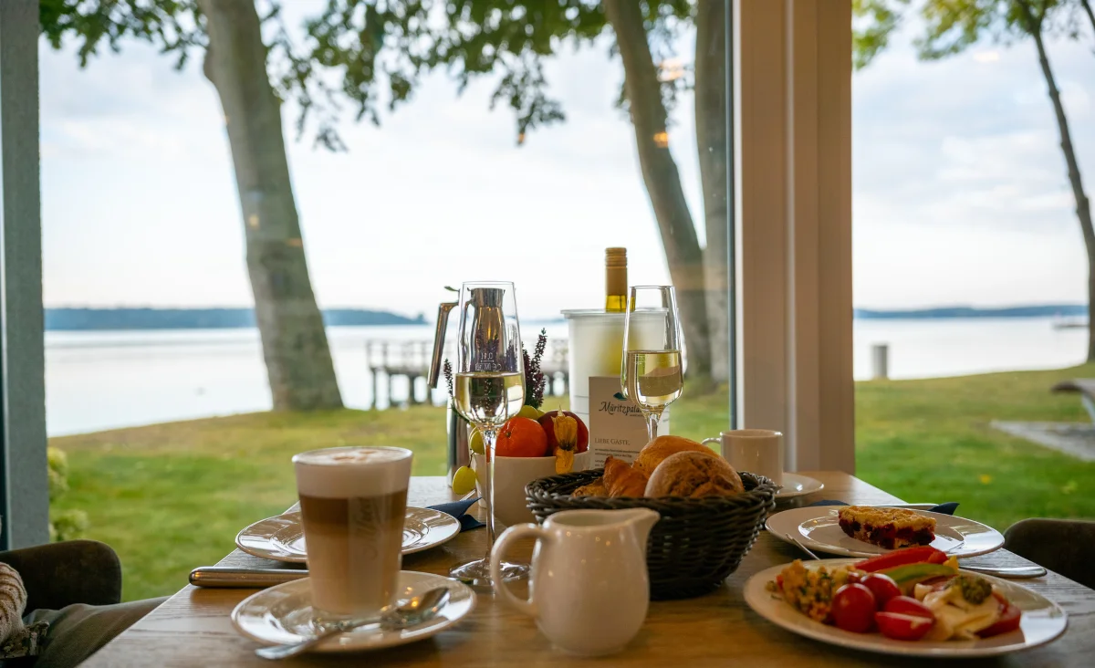 Frühstückstisch mit Kaffee, Croissants, Obst und Wein vor großem Fenster mit Blick auf See und Bäume