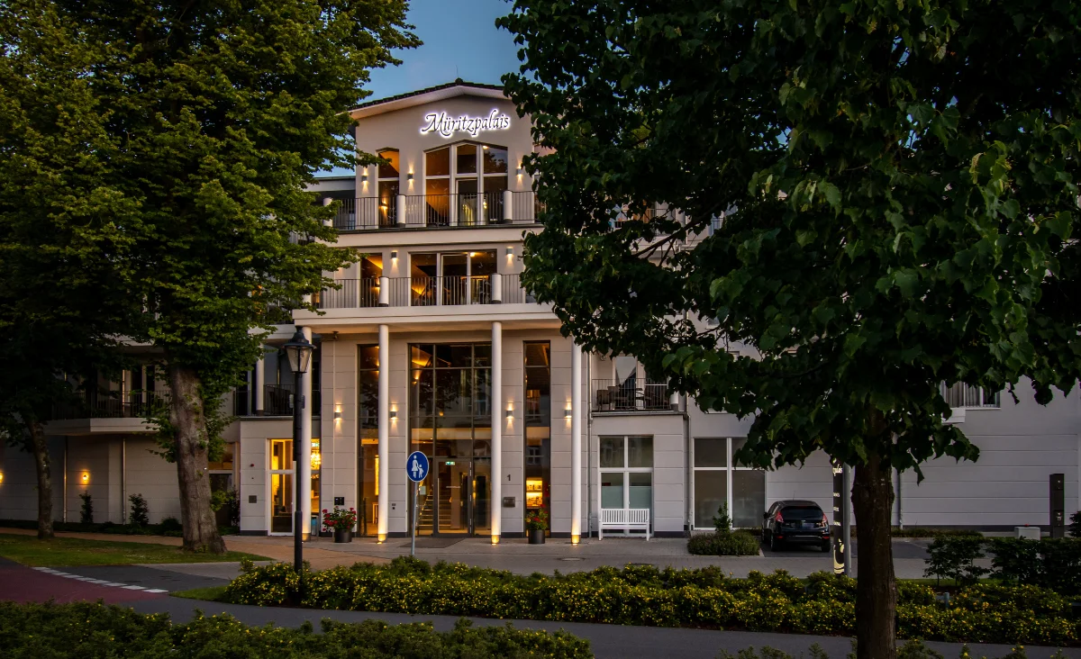 Fassade des Aparthotels Müritzpalais in Waren an der Müritz mit beleuchtetem Schriftzug und umgeben von Bäumen bei Dämmerung