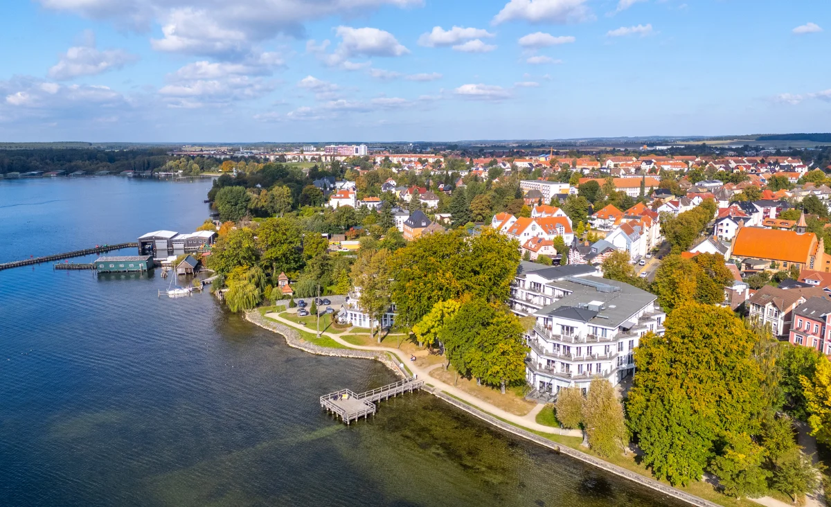 Luftaufnahme des Aparthotels Müritzpalais und der Uferpromenade in Waren an der Müritz mit angrenzendem See, Stegen und umliegenden Gebäuden in einer grünen, städtischen Umgebung