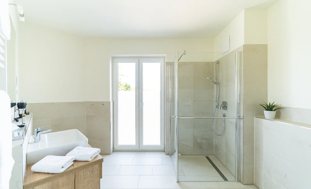 Helles Badezimmer mit bodentiefer Dusche, Doppelwaschbecken auf Holzunterschrank, Handtüchern und Fenster mit Blick nach draußen