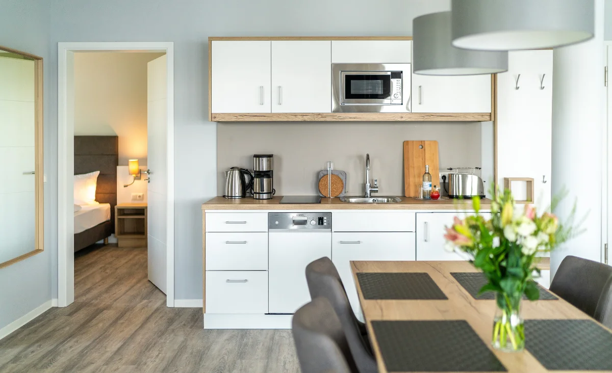 Moderne Küchenzeile mit weißen Schränken, eingebauter Mikrowelle, Kaffeemaschine, Wasserkocher und Esstisch mit grauen Stühlen und Blumenstrauß, im Hintergrund ein offenes Schlafzimmer mit Bett und Nachttischlampe