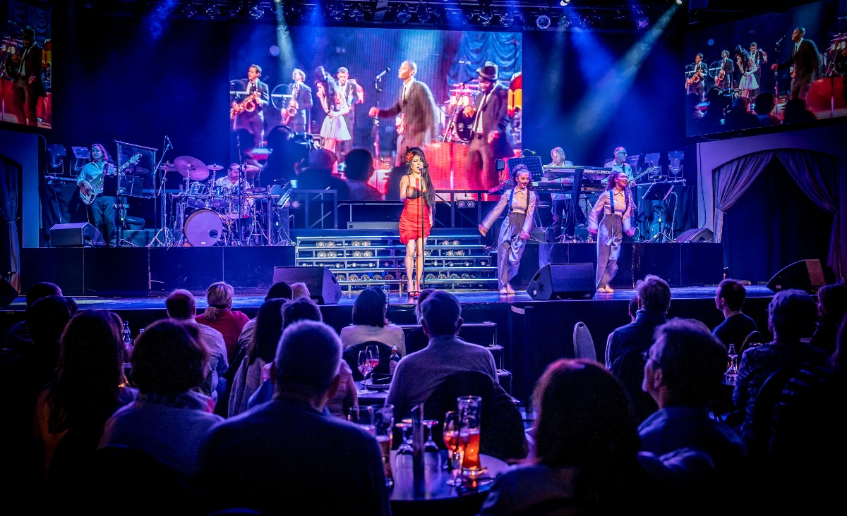 Sängerin in rotem Kleid und schwarzer Perücke mit Hochsteckfrisur steht auf Bühne vor Band und Publikum im Estrel Showtheater Berlin bei Amy Winehouse Tribute Show