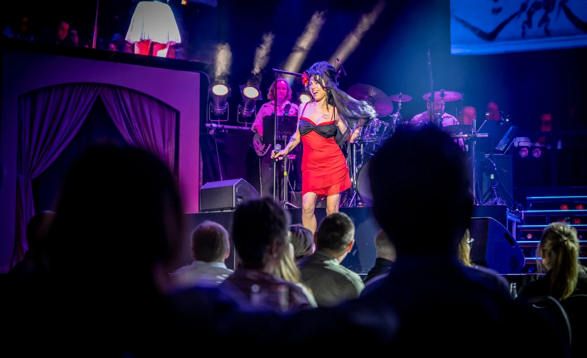 Sängerin in rotem Kleid und schwarzer Perücke mit Hochsteckfrisur steht auf Bühne vor Band und Publikum im Estrel Showtheater Berlin bei Amy Winehouse Tribute Show