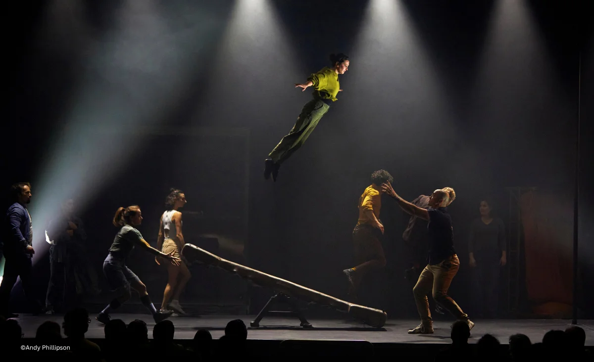 Schauspieler auf der Bühne des Chamäleon Theaters führen eine Szene mit einem Sprung über eine Wippe unter Bühnenbeleuchtung auf