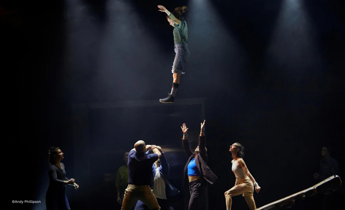 Szenenfoto aus dem Chamäleon Theater mit einer Person, die in der Luft schwebt, während mehrere andere Personen unten die Arme nach oben strecken