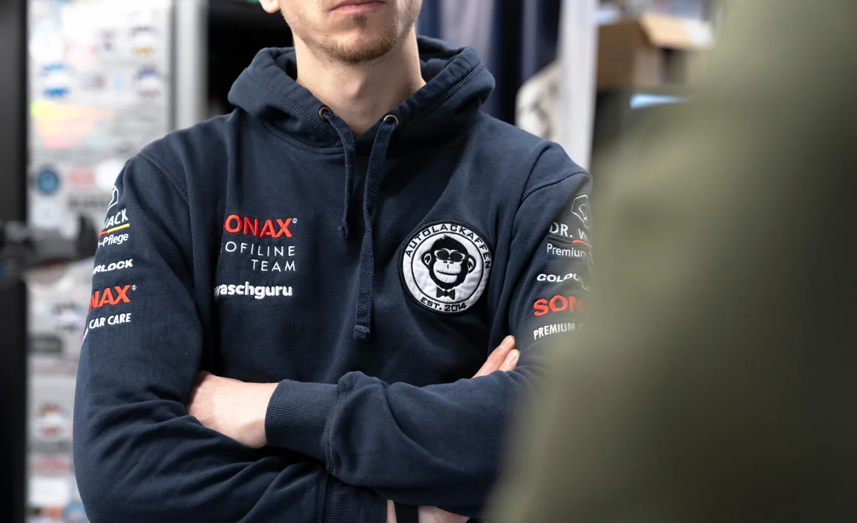 Oberkörper eines Mannes mit verschränkten Armen in einem dunkelblauen Kapuzenpullover mit mehreren Logos, darunter das AUTOLACKAFFEN-Logo mit einem Affengesicht und Schriftzug